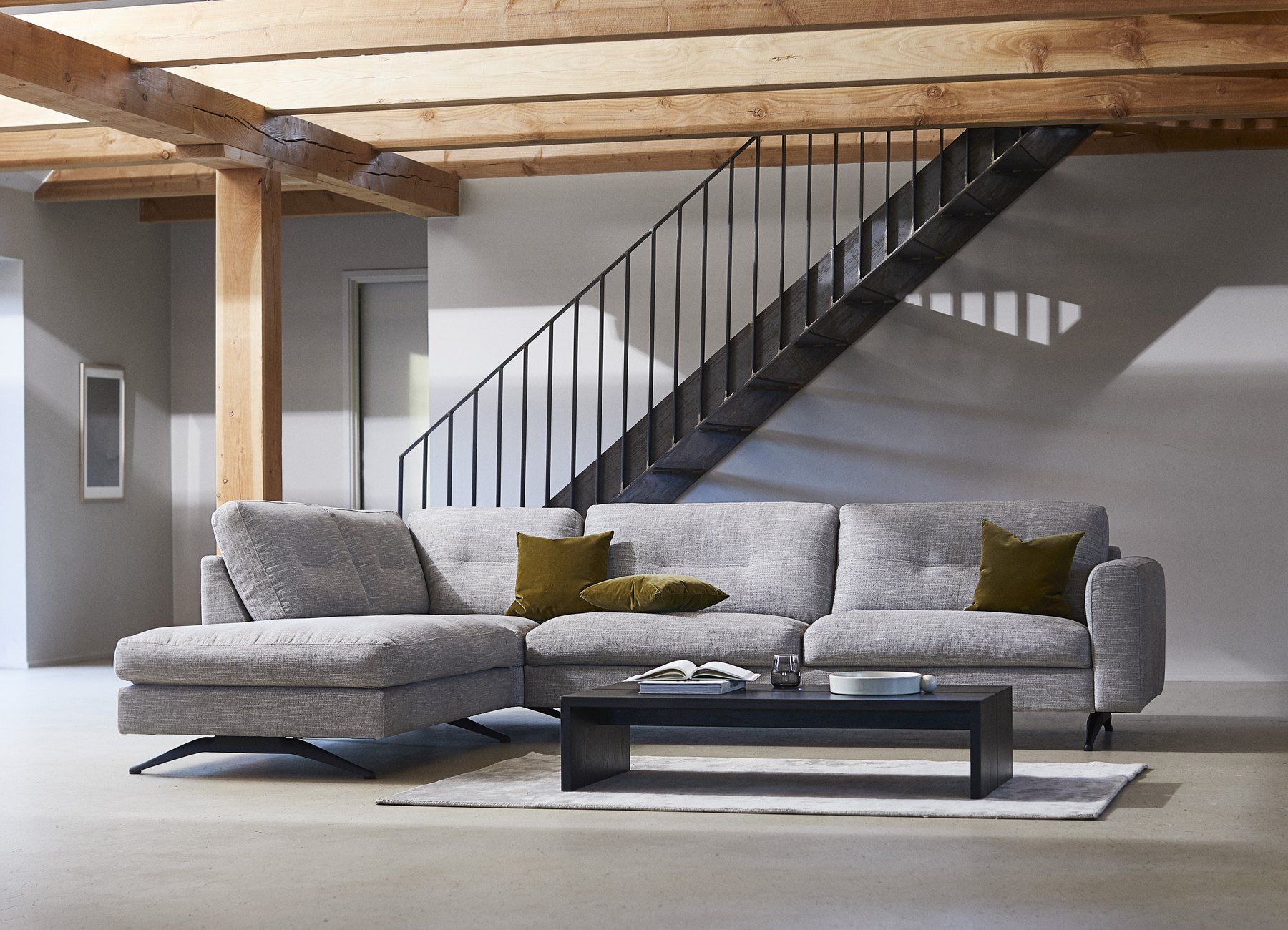 Jasnoszara sofa Flexlux Glow z szezlongiem i oliwkowymi poduszkami, przed nią niski czarny stolik